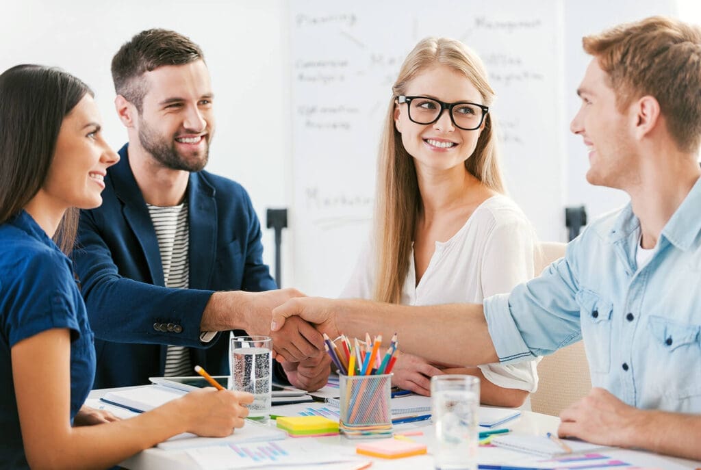 Quatre collègues sont assis autour d'une table dans un bureau, souriant et se serrant la main, avec des documents, des cahiers et des crayons de couleur sur la table et un tableau blanc en arrière-plan.