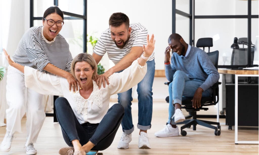 Trois collègues rient et s'amusent tandis qu'une femme monte sur une chaise tandis que deux autres la poussent ; un autre homme est assis en arrière-plan, souriant à la scène ludique dans un espace de bureau moderne.