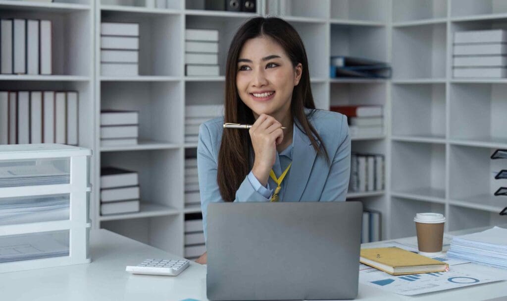 Une femme en blazer bleu clair est assise à un bureau avec un ordinateur portable, un stylo à la main, et sourit. Des étagères remplies de dossiers et de livres occupent l'arrière-plan ; des papiers, une tasse à café et des fournitures de bureau sont posés sur le bureau.