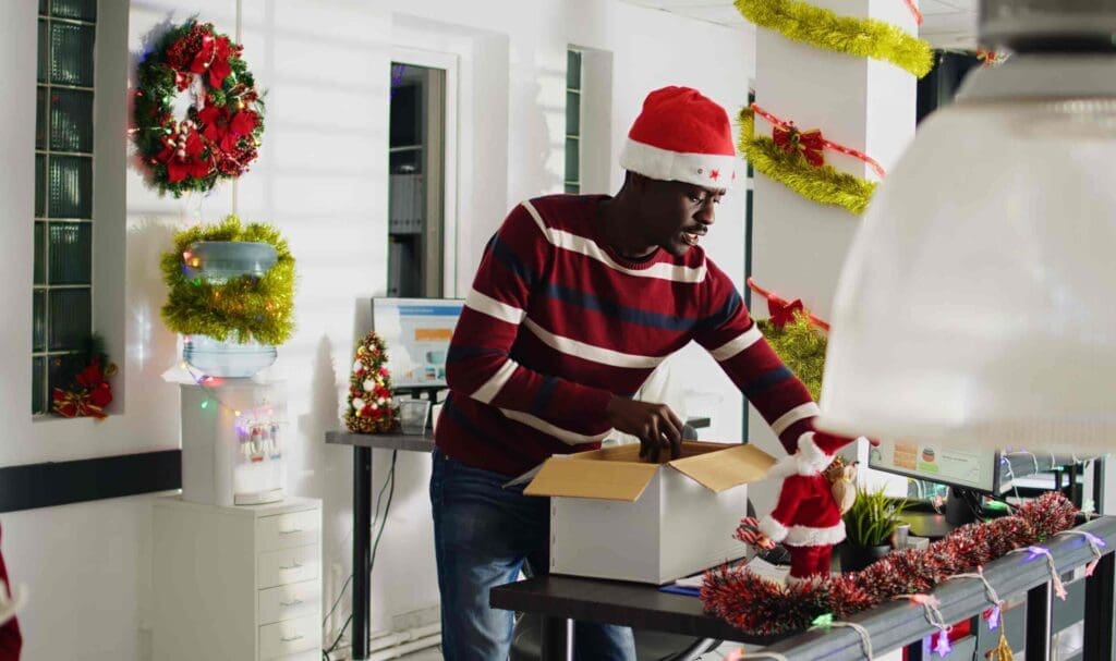 Un homme portant un bonnet de Père Noël et un pull rayé ouvre une boîte en carton sur un bureau décoré, entouré de décorations de Noël, de guirlandes et de décorations festives dans un cadre de bureau.