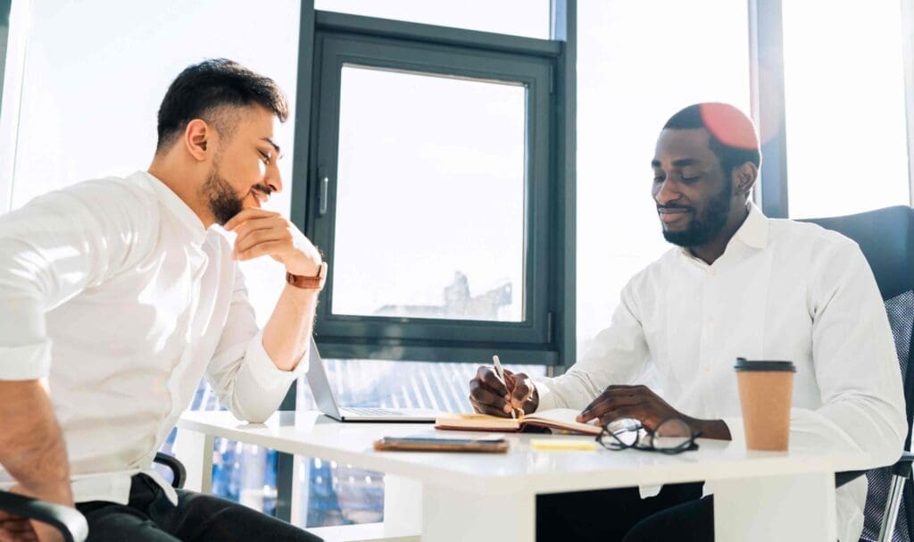 Deux hommes en chemise blanche sont assis à un bureau dans un bureau lumineux. L'un écrit dans un carnet tandis que l'autre regarde pensivement. Un ordinateur portable, un smartphone et une tasse de café sont posés sur le bureau. La lumière du soleil filtre par la fenêtre.