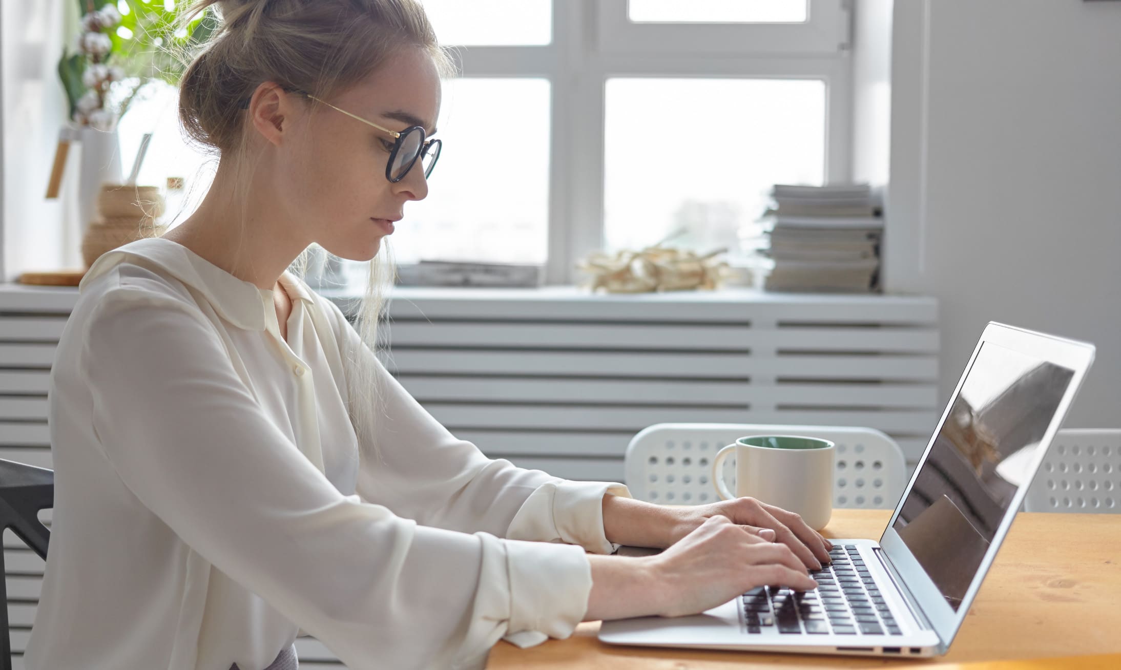 Une femme portant des lunettes et un chemisier blanc est assise à une table et tape sur un ordinateur portable. Une tasse est posée à côté d'elle. Elle travaille dans une pièce lumineuse avec de grandes fenêtres et des piles de papiers en arrière-plan.