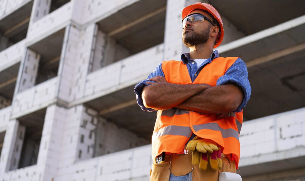 A construction worker wearing an orange safety vest, hard hat, and gloves stands with arms crossed in front of a partially built concrete building, looking confidently into the distance.