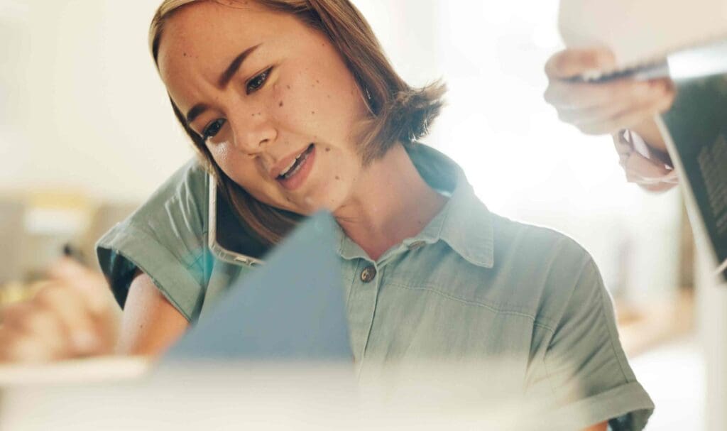 Une femme effectue plusieurs tâches à la fois au travail, tenant son téléphone entre son oreille et son épaule, écrivant sur du papier et consultant les documents qu'on lui tend. Elle semble concentrée et occupée dans un environnement de bureau.