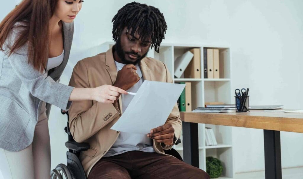 Une femme se tient à côté d'un homme assis en fauteuil roulant, tous deux regardant un document. La femme pointe le papier du doigt tandis que l'homme le regarde pensivement. Ils se trouvent dans un bureau moderne.
