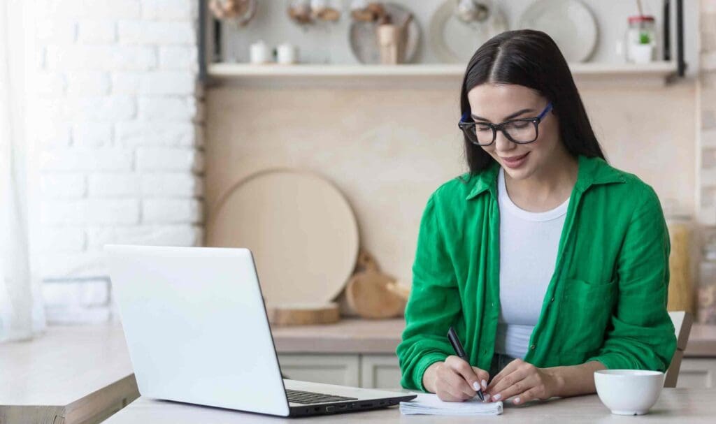 Une femme portant des lunettes et une chemise verte est assise à une table, écrivant dans un cahier avec un ordinateur portable et une tasse à proximité, dans une cuisine lumineuse et confortable.