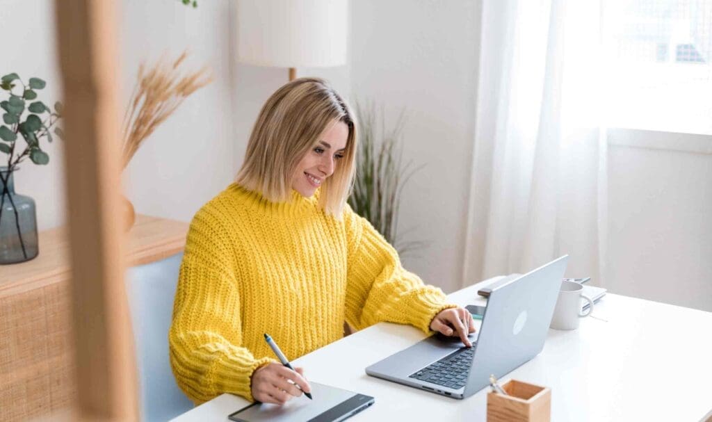Une femme vêtue d'un pull jaune est assise à un bureau, souriante, travaillant sur un ordinateur portable et écrivant sur un bloc-notes. Le bureau est équipé d'une tasse, d'un pot à crayons et de plantes, et la lumière naturelle provient d'une fenêtre voisine.