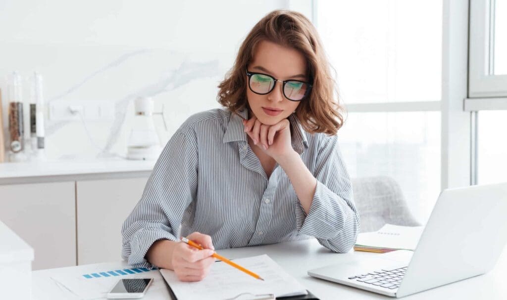 Une femme portant des lunettes est assise à un bureau et écrit sur un document au crayon. Elle est entourée d'un ordinateur portable, de graphiques et d'un smartphone, semblant concentrée et réfléchie dans un bureau moderne et lumineux.