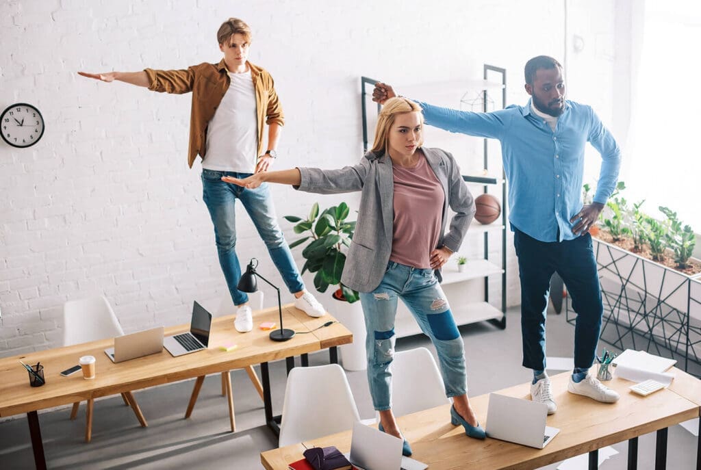Trois personnes se tiennent debout sur des bureaux dans un bureau lumineux, affichant une posture assurée, digne d'un super-héros, un bras tendu. Ordinateurs portables, carnets et plantes sont visibles sur les bureaux.
