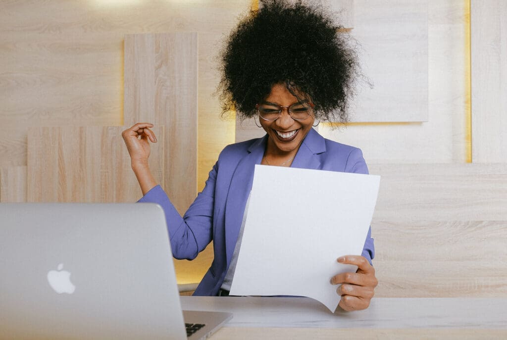 Une femme portant des lunettes et un blazer violet sourit et tient une feuille de papier tout en étant assise à un bureau avec un ordinateur portable ouvert, semblant heureuse et excitée.
