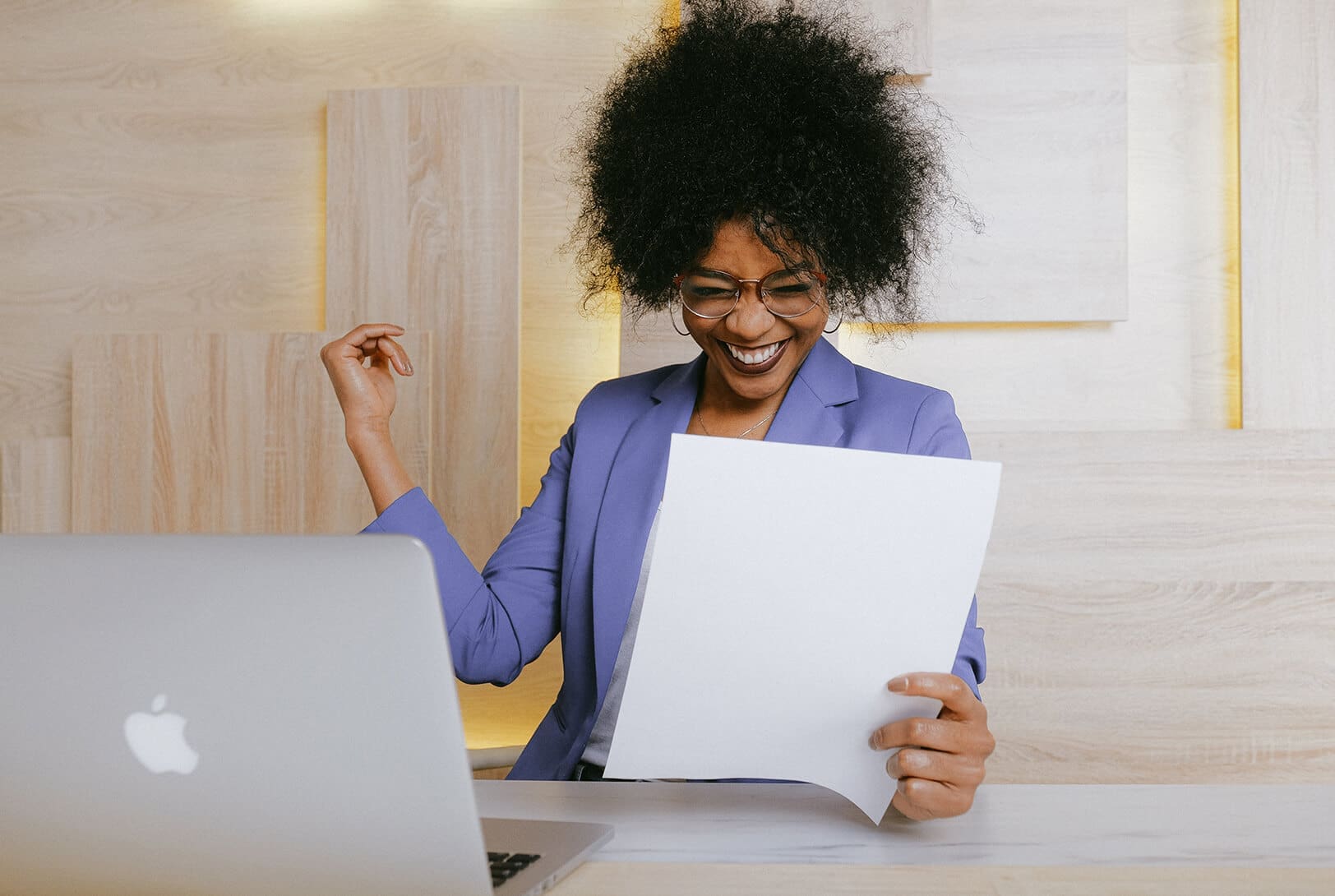 Une femme portant des lunettes et un blazer violet sourit et tient une feuille de papier tout en étant assise à un bureau avec un ordinateur portable ouvert, semblant heureuse et excitée.