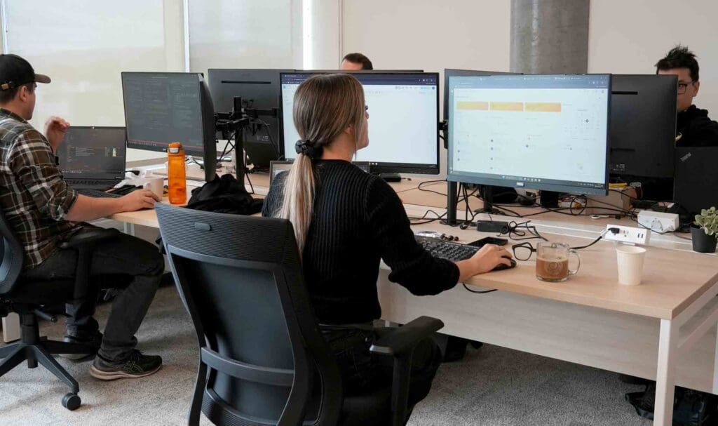 Four people work at desks with large computer monitors in a modern office. One woman sits in the foreground, typing, with a coffee mug and water bottle nearby. The room has bright windows and a casual atmosphere.