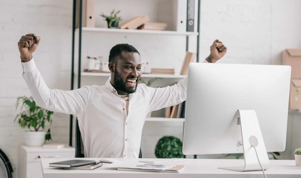 A man sitting at a desk in front of a computer, smiling and raising both arms in celebration, in a bright, modern office with shelves and plants in the background.