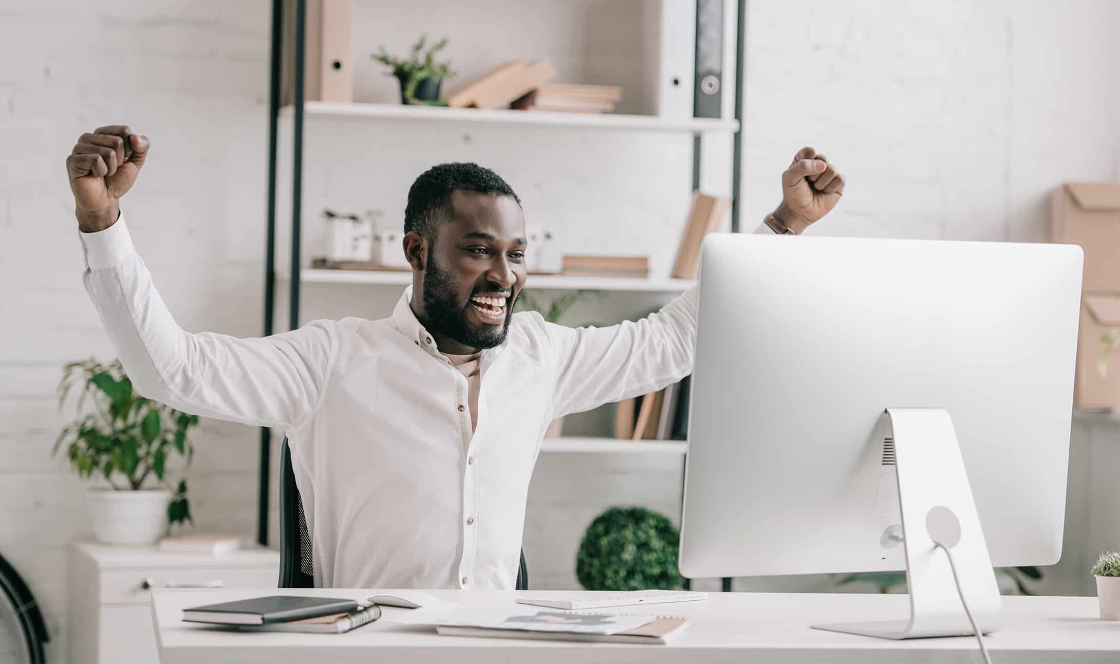 A man sitting at a desk in front of a computer, smiling and raising both arms in celebration, in a bright, modern office with shelves and plants in the background.