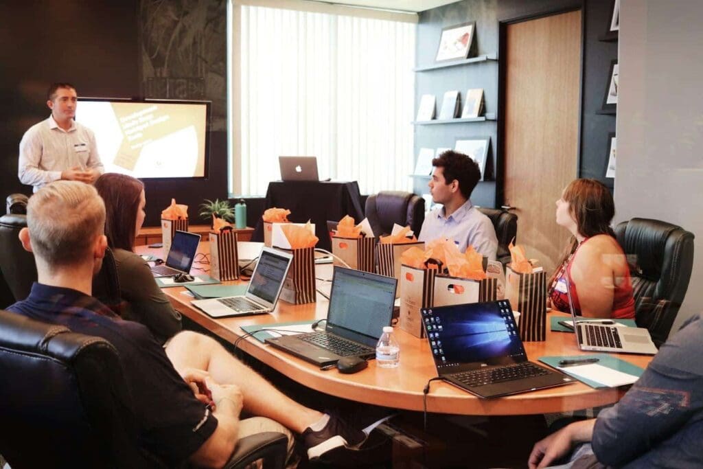 A group of people sit around a conference table with laptops and gift bags, listening to a man giving a presentation in a modern office meeting room.