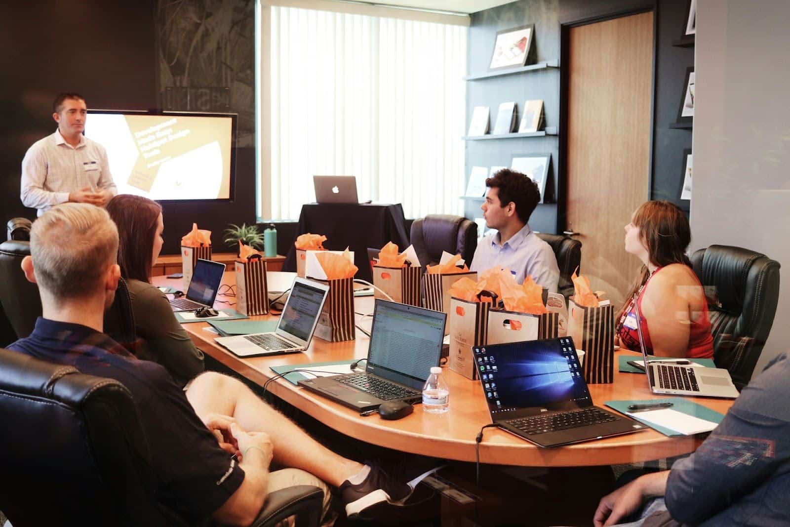 A group of people sit around a conference table with laptops and gift bags, listening to a man giving a presentation in a modern office meeting room.