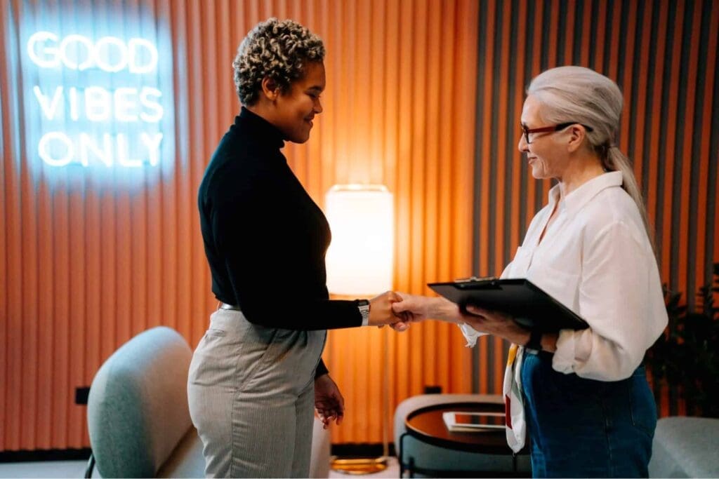 Deux femmes se serrent la main et sourient dans un bureau moderne avec un néon « GOOD VIBES ONLY » et un éclairage chaleureux en arrière-plan. L'une d'elles tient un presse-papiers.