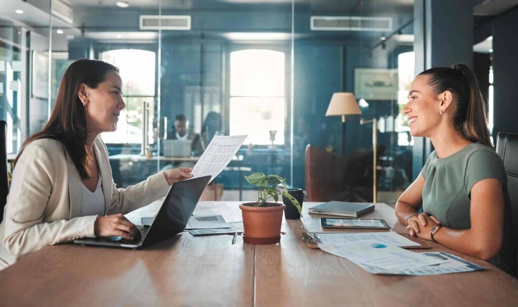 Deux femmes sont assises face à face à une table de bureau, souriantes et discutant. L'une tient un document, l'autre écoute. Des papiers, un ordinateur portable et une plante en pot sont posés sur la table. Le bureau est vitré.