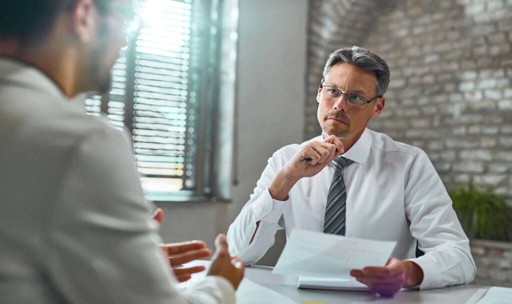 Un homme en chemise blanche et cravate est assis à un bureau, des papiers à la main, et écoute attentivement une personne qui parle en gestuelle. La lumière du soleil filtre à travers les stores d'un bureau moderne avec un mur de briques.