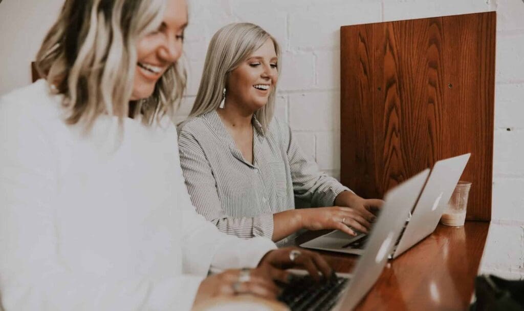 Two women sit side by side at a wooden desk, smiling and working on laptops. A coffee cup is placed next to one of them. The background features white brick walls and wooden dividers.