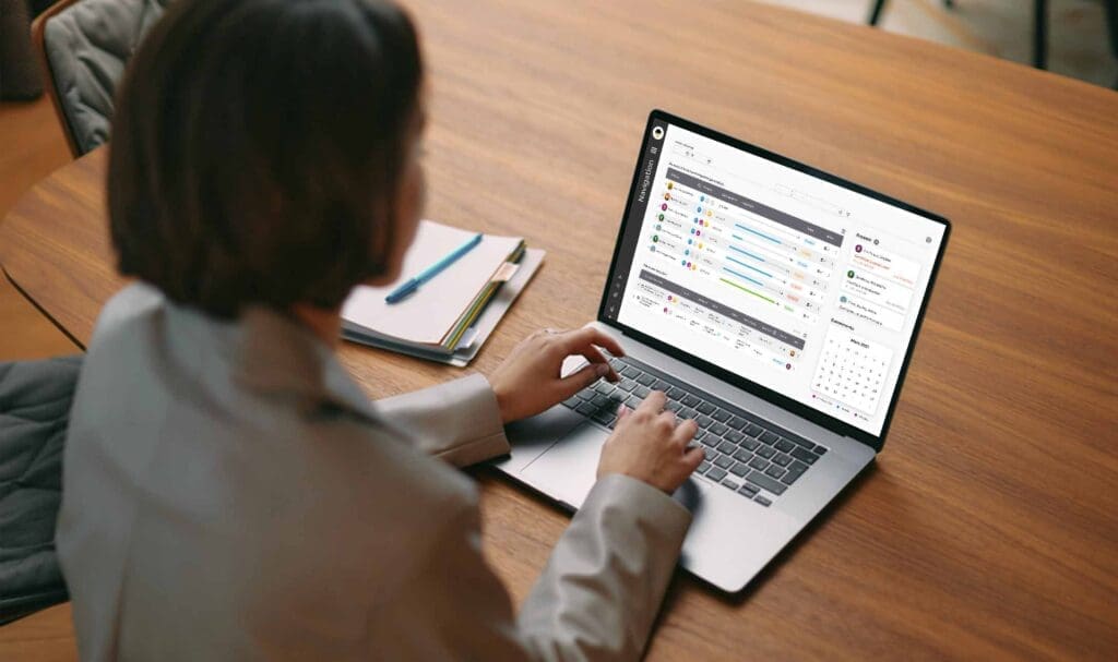 Une personne en blazer travaille sur un ordinateur portable, assis sur une table en bois, avec une application de gestion de projet ou de planification comportant des listes de tâches, des barres de progression et un calendrier. Des cahiers et un stylo sont à proximité.