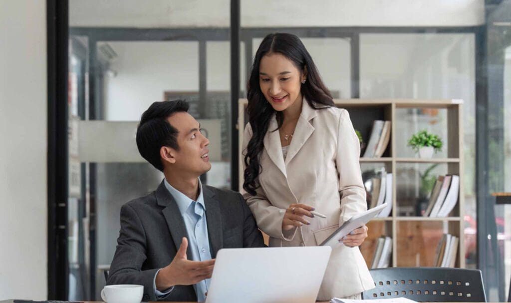 Un homme assis à un bureau parle à une femme debout à côté de lui, tous deux en tenue professionnelle, dans un bureau moderne avec un ordinateur portable, des documents et des étagères en arrière-plan.