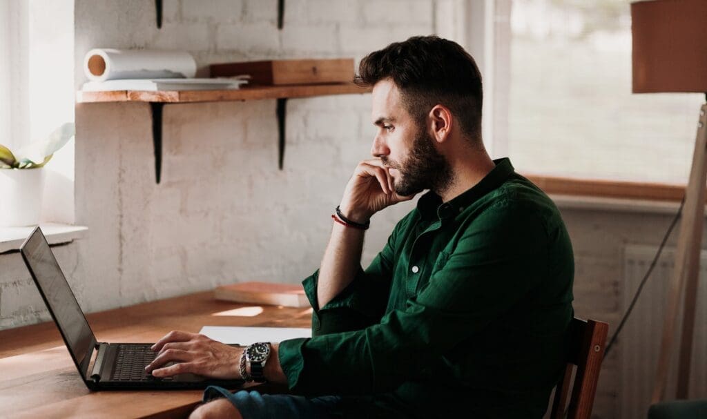 Un homme barbu est assis à un bureau en bois, concentré sur son ordinateur portable. Il porte une chemise verte et pose son menton sur sa main. Des étagères et une lampe sont visibles dans la pièce lumineuse et chaleureuse.
