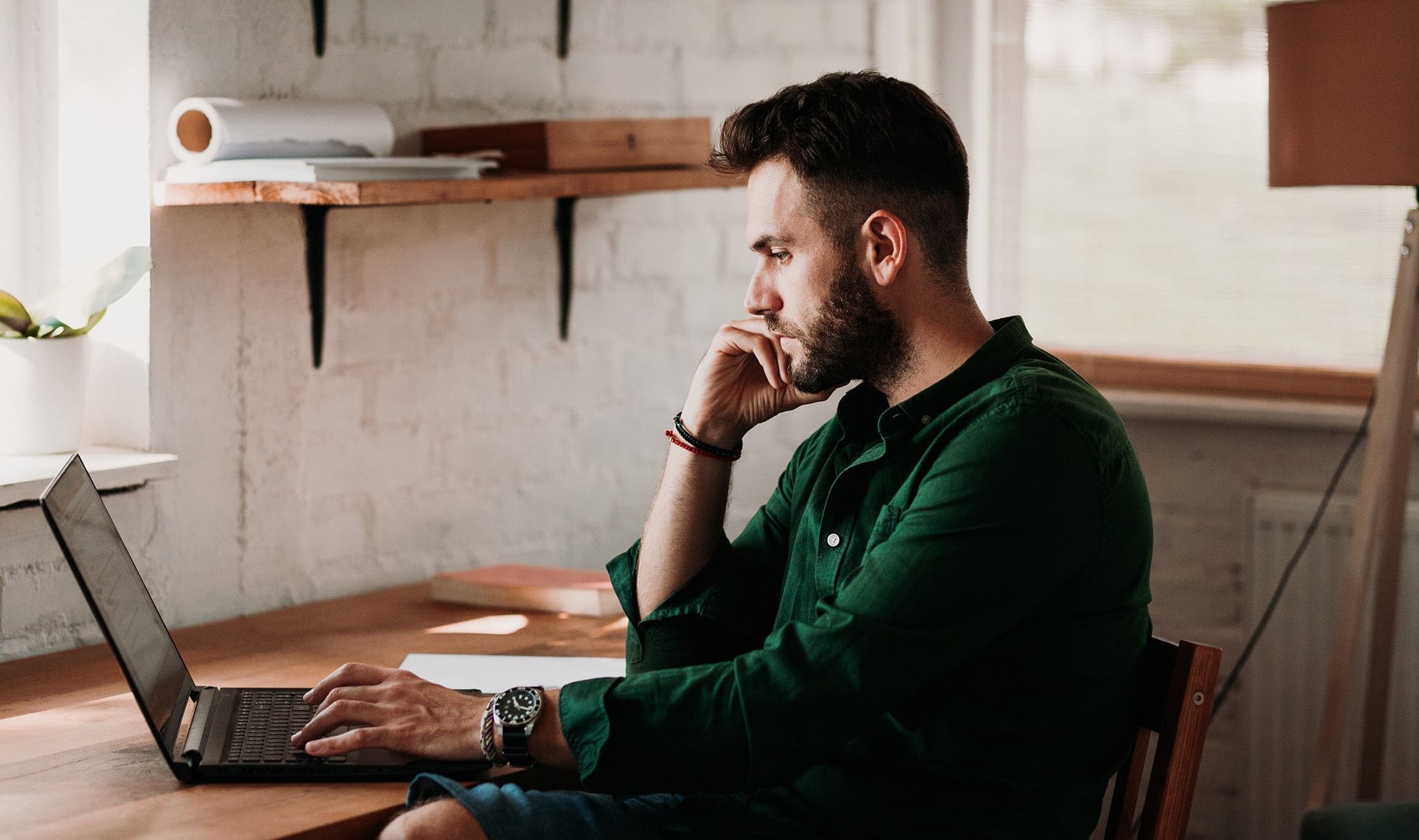 Un homme barbu est assis à un bureau en bois, concentré sur son ordinateur portable. Il porte une chemise verte et pose son menton sur sa main. Des étagères et une lampe sont visibles dans la pièce lumineuse et chaleureuse.