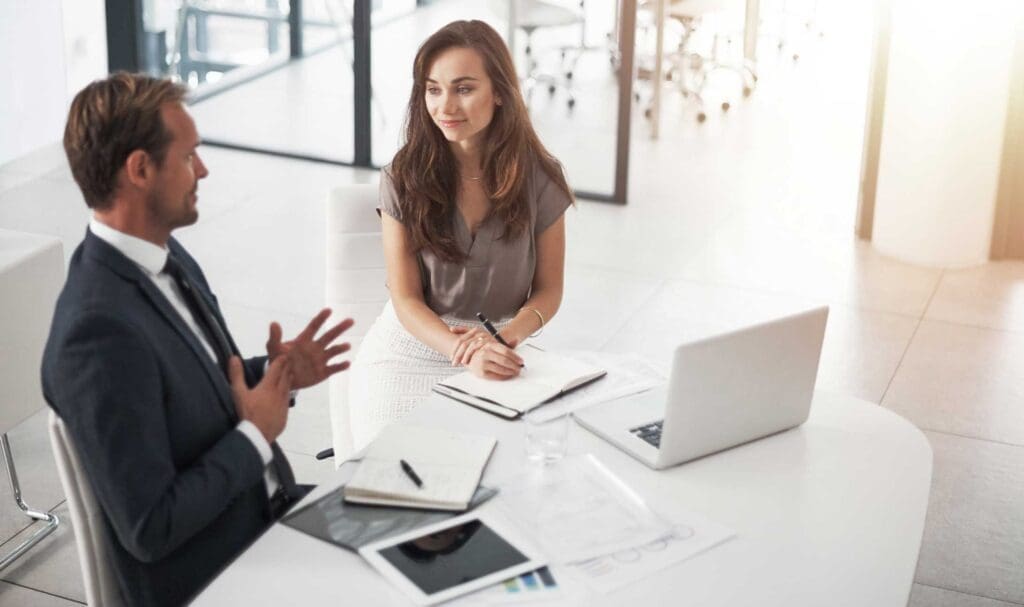 Un homme en costume et une femme en tenue professionnelle sont assis à un bureau blanc dans un bureau moderne, discutant et prenant des notes. Des papiers, un ordinateur portable et une tablette sont posés sur le bureau. L'ambiance est professionnelle et collaborative.