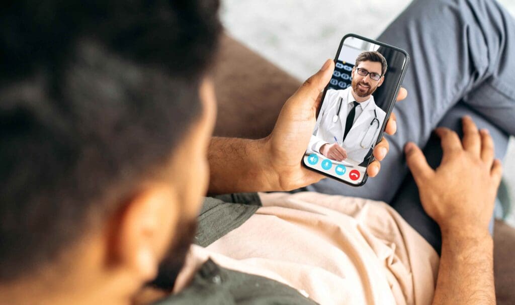 Une personne tient un smartphone et participe à un appel vidéo avec un médecin portant une blouse blanche et un stéthoscope. La conversation semble amicale et professionnelle.