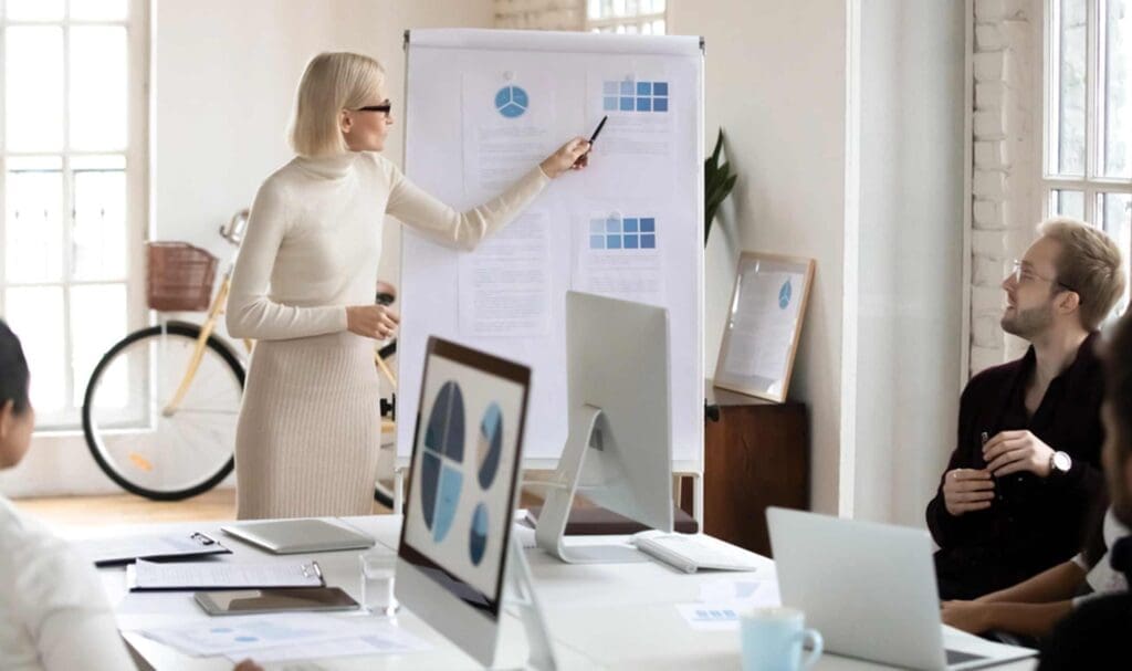 Dans un bureau moderne, une femme vêtue d'un pull léger présente des graphiques sur un tableau à ses collègues. Des ordinateurs portables et des documents sont posés sur la table, et un vélo est visible en arrière-plan.