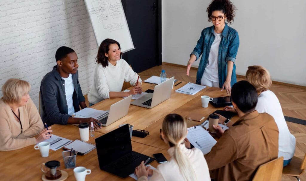 Un groupe de six personnes est assis autour d'une table avec des ordinateurs portables et des papiers, tandis qu'une personne se tient debout et fait des gestes, menant une discussion dans un cadre de bureau lumineux et moderne.