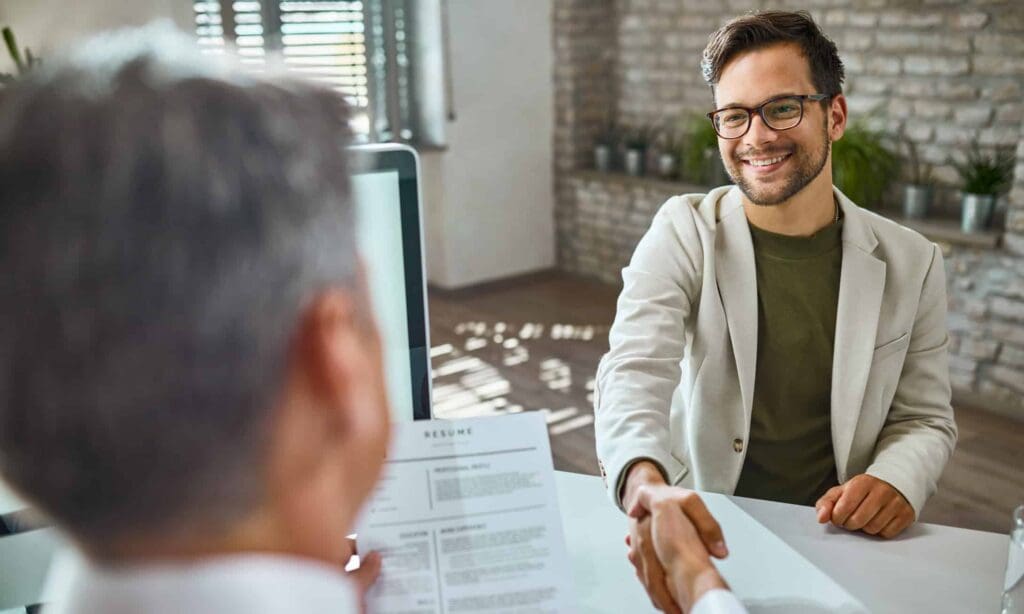 Un jeune homme souriant, portant des lunettes et un blazer beige, serre la main d'une autre personne assise à un bureau lors d'un entretien d'embauche. Un CV est visible au premier plan, et le décor semble être un bureau.