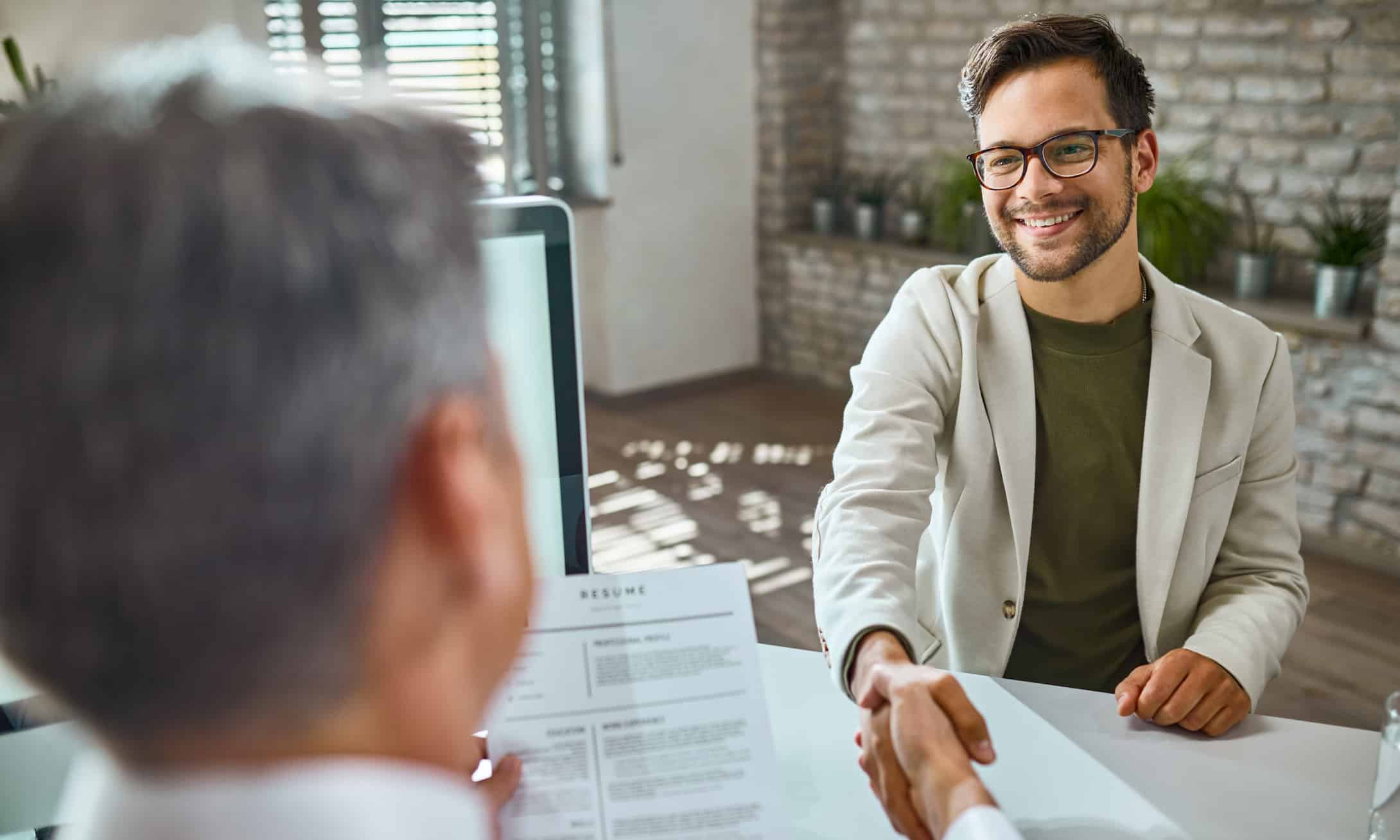 Un jeune homme souriant, portant des lunettes et un blazer beige, serre la main d'une autre personne assise à un bureau lors d'un entretien d'embauche. Un CV est visible au premier plan, et le décor semble être un bureau.