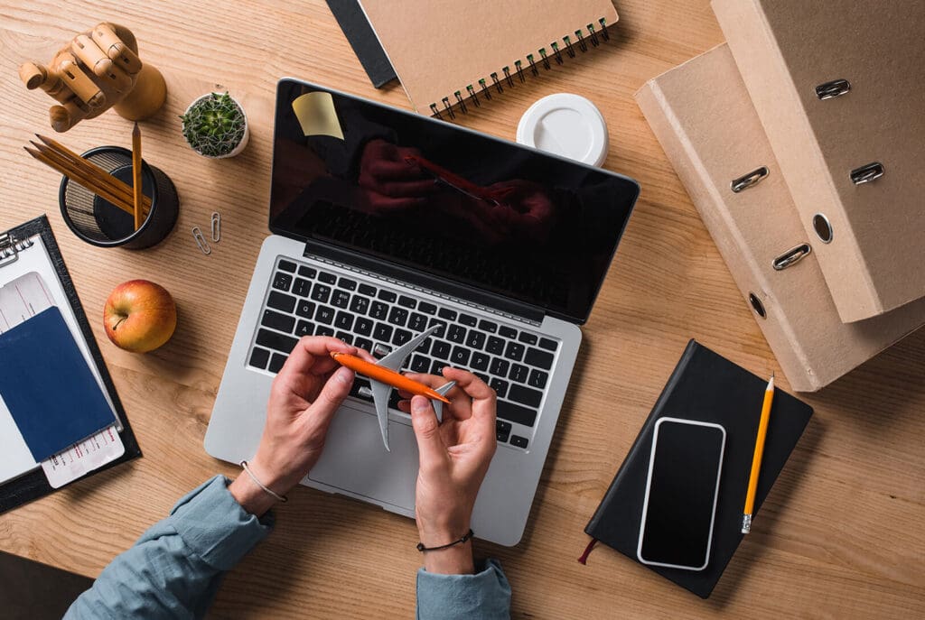 Vue de dessus d'une personne assise à un bureau avec un ordinateur portable, un stylo orange à la main. Le bureau contient des dossiers, des cahiers, un smartphone, une pomme, une tasse à café, une plante et des fournitures de bureau.