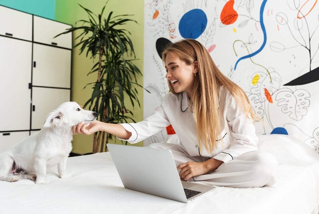 Une jeune femme en pyjama est assise sur un lit avec un ordinateur portable, souriante, tout en caressant doucement un petit chien blanc. La pièce est décorée d'œuvres d'art murales colorées et abstraites, avec une plante verte en arrière-plan.