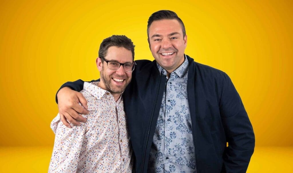 Deux hommes souriants, debout l'un près de l'autre devant un fond jaune vif. L'un d'eux passe son bras autour des épaules de l'autre. Tous deux portent des chemises à motifs et des lunettes.