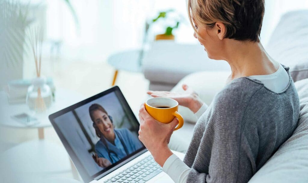 A person holding a yellow mug sits on a couch and has a video call with a smiling woman on a laptop screen in a bright, modern living room.