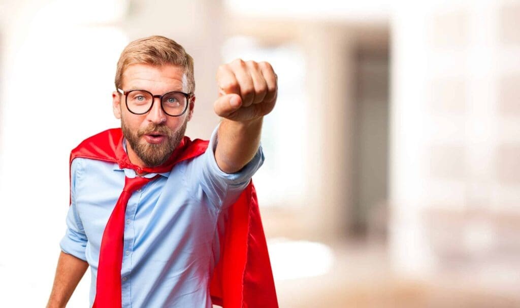 Un homme portant des lunettes, une cape et une cravate rouges pose comme un super-héros, le poing levé comme s'il volait. Il a l'air confiant et porte une chemise bleu clair. L'arrière-plan est flou.