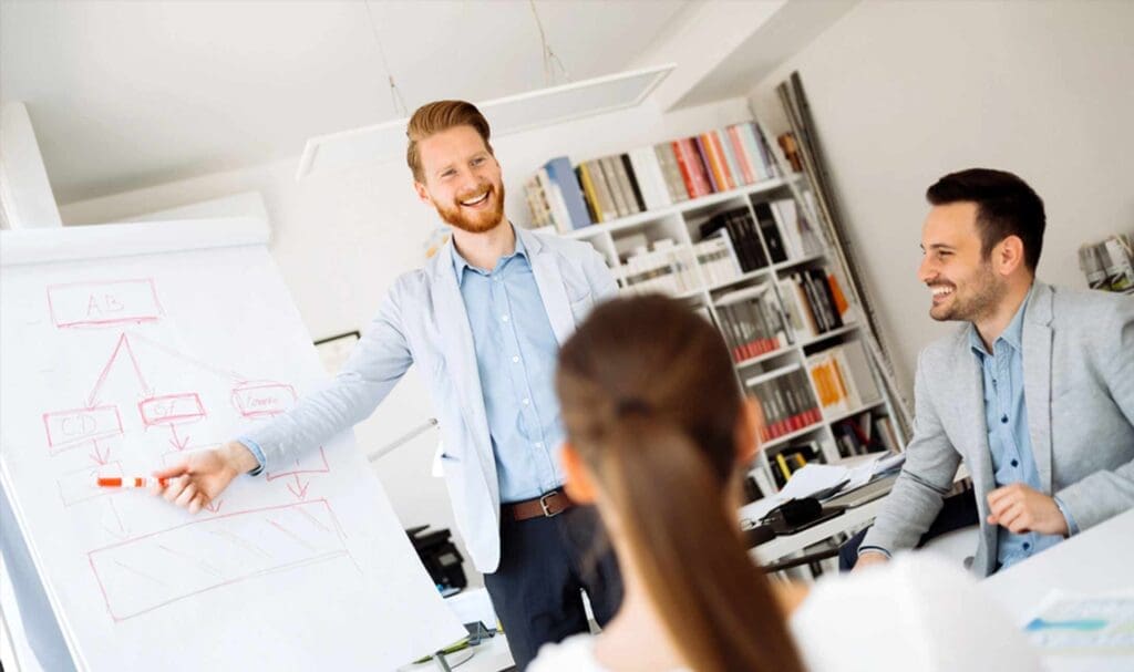 Un homme se tient devant un tableau de conférence, souriant et pointant un diagramme du doigt lors d'une réunion. Deux collègues, un homme et une femme, sont assis à la table, écoutant et souriant dans un bureau lumineux et moderne.