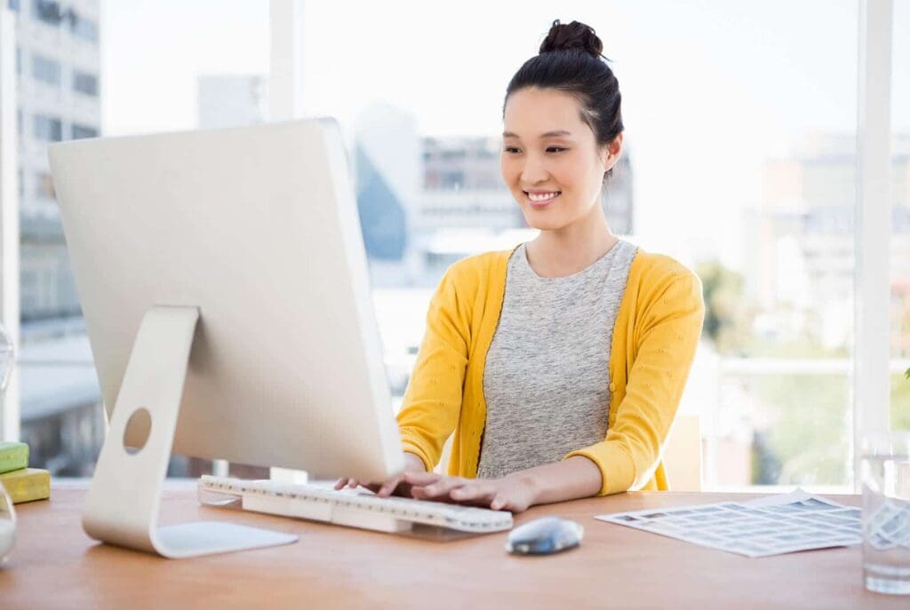 A woman wearing a yellow cardigan and gray shirt smiles while working at a desktop computer in a bright office with large windows. Papers and a mouse are on the wooden desk.