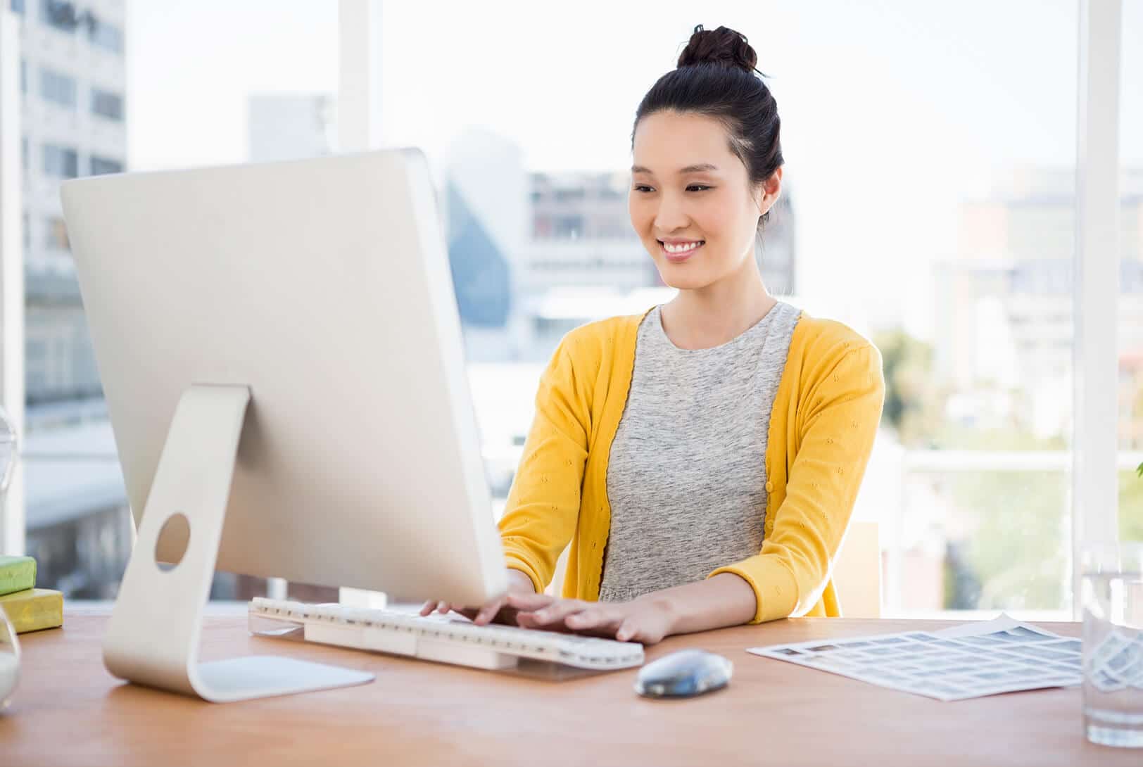 A woman wearing a yellow cardigan and gray shirt smiles while working at a desktop computer in a bright office with large windows. Papers and a mouse are on the wooden desk.