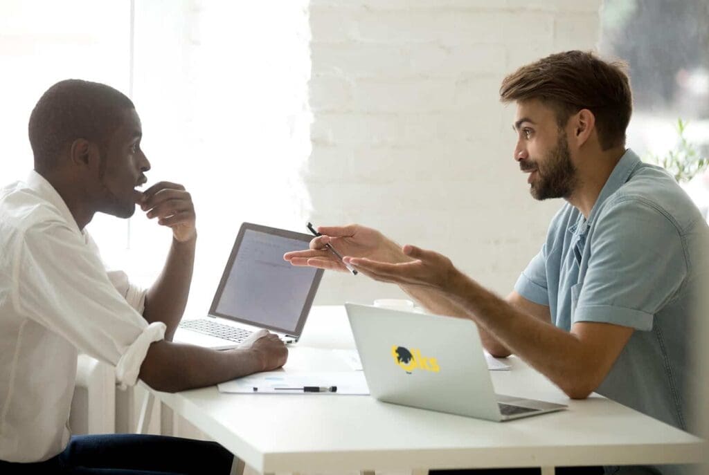 Deux hommes sont assis face à face à un bureau avec des ordinateurs portables, en pleine conversation. L'un fait des gestes avec un stylo, tandis que l'autre écoute pensivement, la main sur le menton.