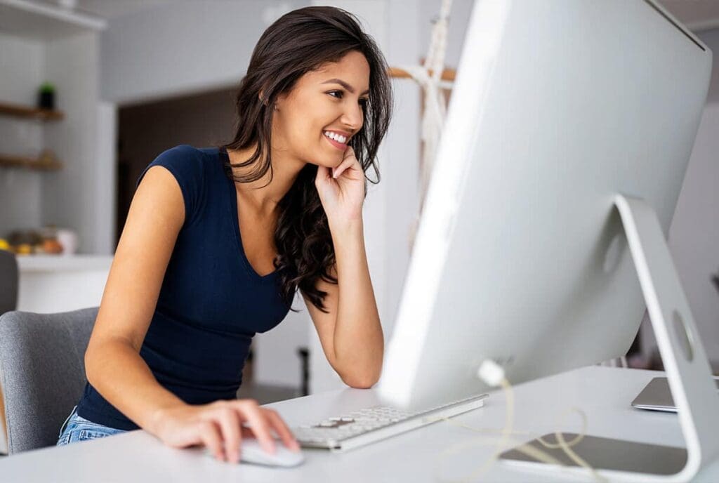 Une femme aux longs cheveux noirs est assise à un bureau, souriante devant un ordinateur. Elle porte une chemise bleu marine et semble travailler dans un bureau à domicile lumineux et moderne.