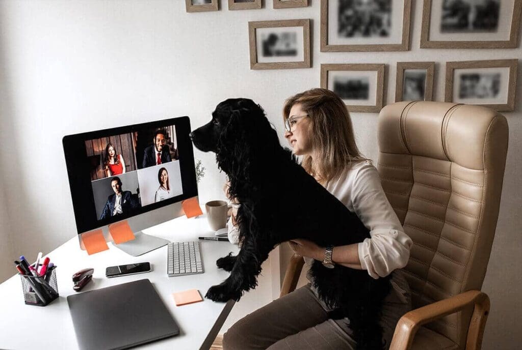 Une femme est assise à un bureau, un chien noir sur les genoux, et regarde un écran d'ordinateur diffusant un appel vidéo avec quatre personnes. Des fournitures de bureau et une tasse à café sont disposées sur le bureau, tandis que des photos encadrées sont accrochées au mur derrière elle.