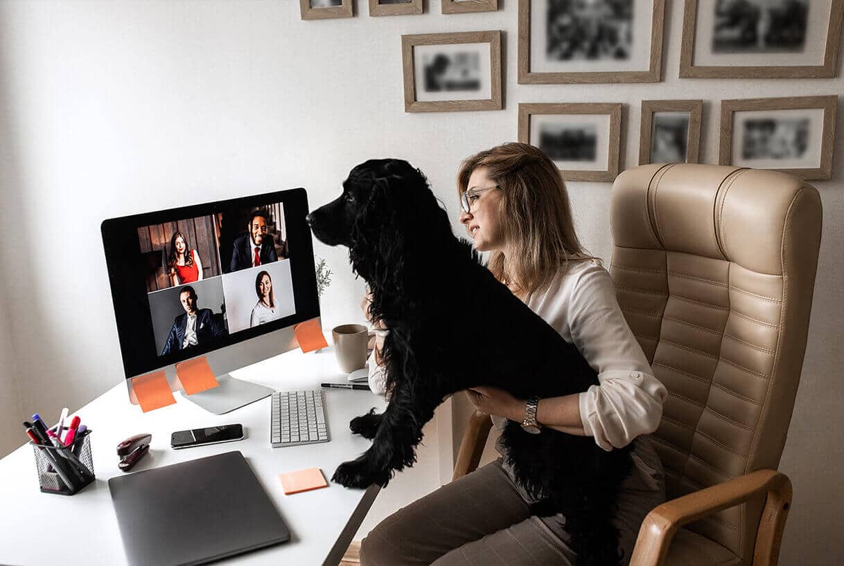 Une femme est assise à un bureau, un chien noir sur les genoux, et regarde un écran d'ordinateur diffusant un appel vidéo avec quatre personnes. Des fournitures de bureau et une tasse à café sont disposées sur le bureau, tandis que des photos encadrées sont accrochées au mur derrière elle.