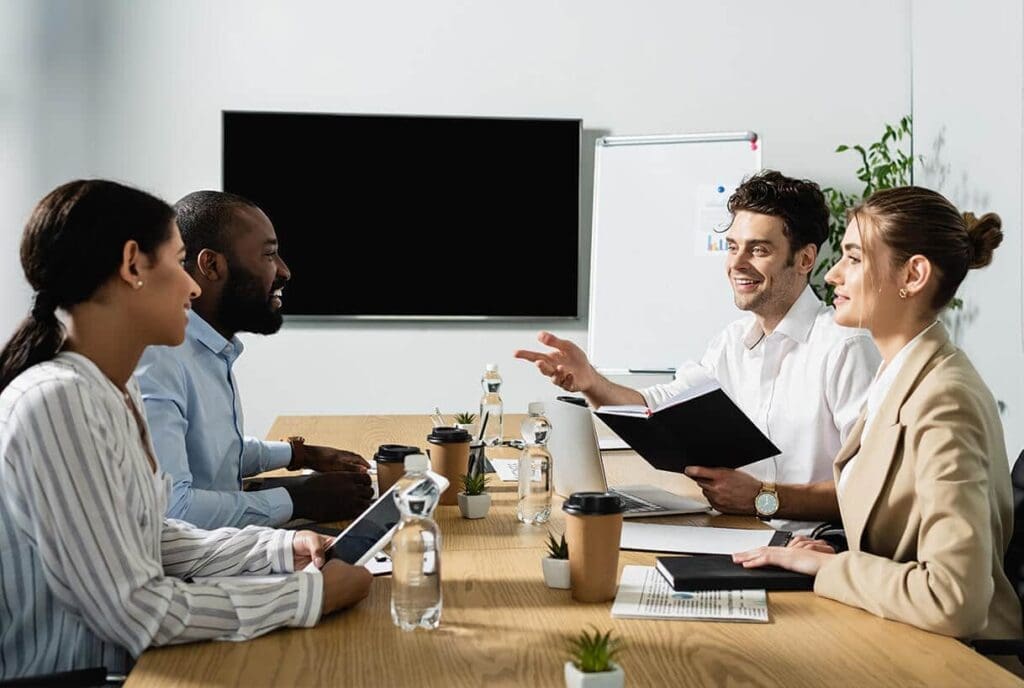Quatre professionnels sont assis autour d'une table de conférence et discutent. Ils sourient, tenant des cahiers et des tablettes. Des bouteilles d'eau, des tasses à café et des plantes sont disposées sur la table, avec un écran de télévision vierge et un tableau de conférence en arrière-plan.