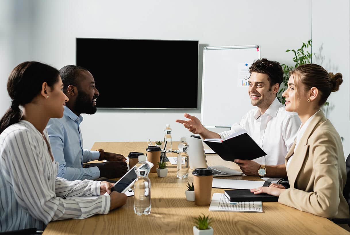Quatre professionnels sont assis autour d'une table de conférence et discutent. Ils sourient, tenant des cahiers et des tablettes. Des bouteilles d'eau, des tasses à café et des plantes sont disposées sur la table, avec un écran de télévision vierge et un tableau de conférence en arrière-plan.