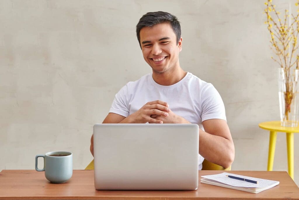 Une personne souriante en t-shirt blanc est assise à un bureau avec un ordinateur portable, une tasse à café, un bloc-notes et un stylo. Une table jaune avec un vase de fleurs se détache sur un mur clair.