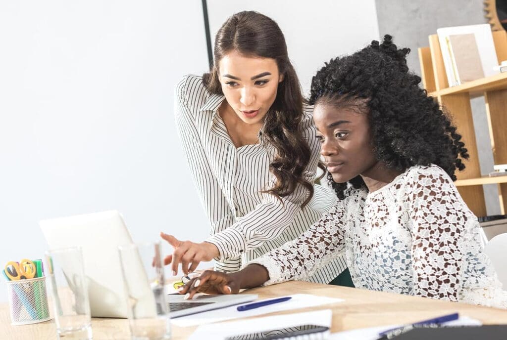 Deux femmes travaillent ensemble à un bureau, les yeux rivés sur l'écran d'un ordinateur portable. L'une est assise et tape du texte, tandis que l'autre se tient à côté d'elle, pointant l'écran du doigt et expliquant quelque chose. Des fournitures de bureau sont visibles sur le bureau.
